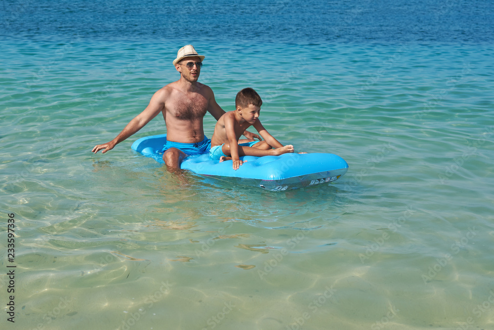 Dad and son are sitting on a blue inflatable floater in the clear ocean water. They are having fun and enjoying their vacations.