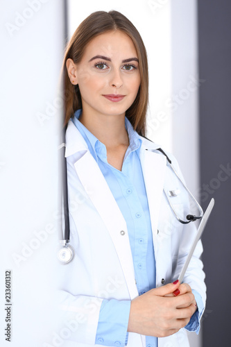 Doctor woman using tablet computer while standing straight near window in hospital. Happy physician at work. Medicine and health care concept