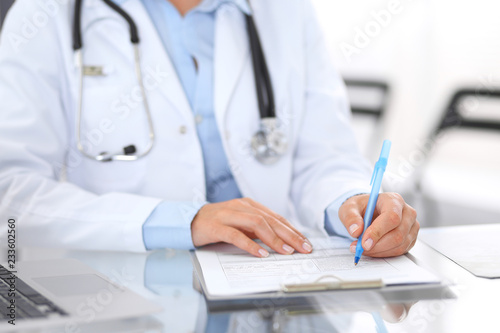 Unknown doctor woman at work  hands close-up. Female physician filling up medical form while sitting at glass desk at hospital office. Medicine and healthcare concept