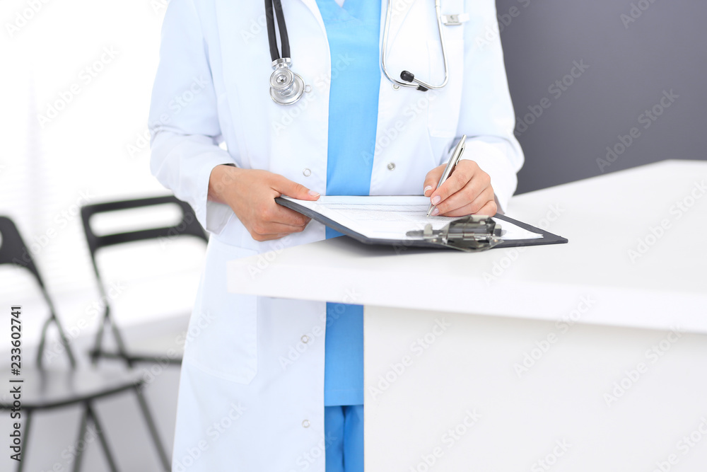 Unknown doctor woman at work, hands close-up. Female physician filling up medical form while standing near reception desk at clinic or emergency hospital. Medicine and healthcare concept Unknown doctor woman at work, hands close-up. Female physician filling up medical form while standing near reception desk at clinic or emergency hospital. Medicine and healthcare concept
