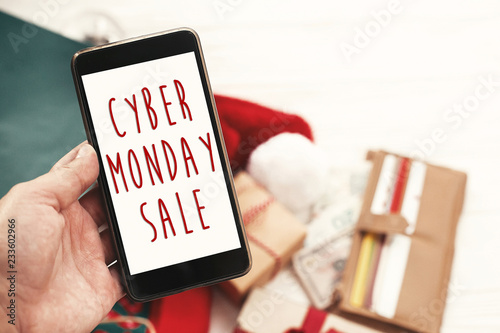 Cyber Monday sale text on phone screen, xmas sale sign. Special discount christmas offer sign. Hand holding phone with advertising message at credit cards, bags, clothes, gifts
