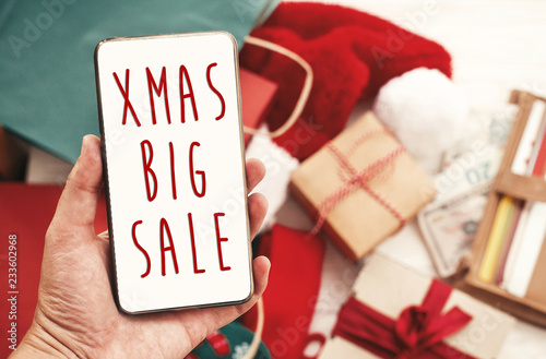 Christmas big sale text on phone screen, xmas sale sign. Special discount christmas offer sign. Hand holding phone with advertising message at credit cards, bags, clothes, gifts