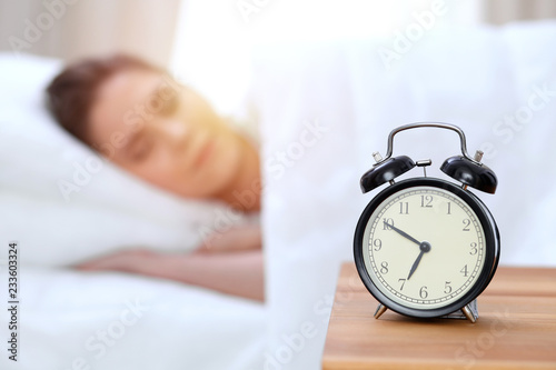 Alarm clock opposite of beautiful young woman sleeping while lying in bed comfortably and blissfully. Happy waking up early for a job