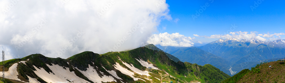 panorama of the mountains and Aibga Ridge with low clouds. Remnants of snow and new green grass on Mountains near the ski resort of Rosa Khutor in Krasnaya Polyana. Sochi, Russia.