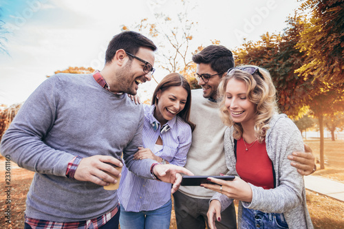 Four happy smiling young friends walking outdoors in the park holding digital tablet
