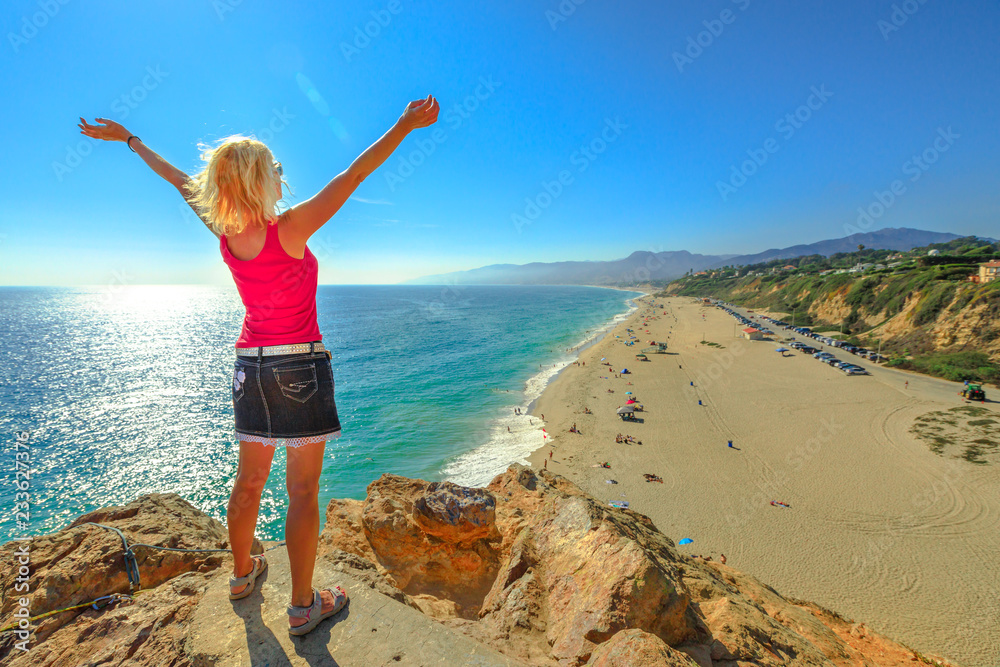 Caucasian female looking Point Dume State Beach from Point Dume promontory on Malibu coast in CA, United States. Carefree woman with open arms in California West Coast. Blue sky, sunny. Copy space.