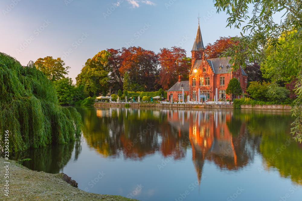 Scenic city view of Lake Minnewater and beautiful medieval house with its reflection at the evening golden hour, Bruges, Belgium