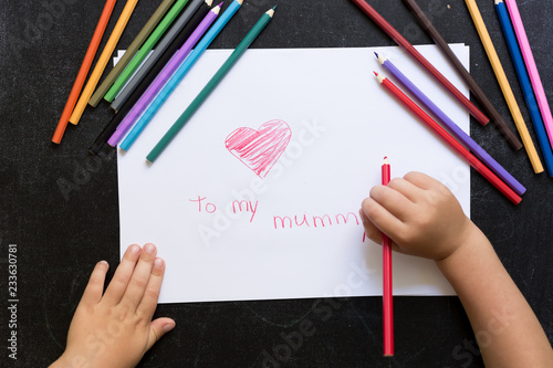 Kid's hands with pencil draws heart on white paper. Mother's day concept. Celebration. Hand made card. Top view. To my mummy words.