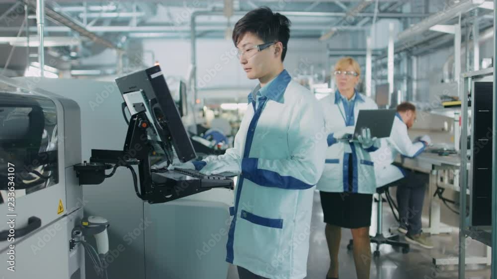 On High Tech Factory Asian Engineer Uses Computer for Programing Pick and Place Electronic Machinery for Printed Circuit Board Surface Mount Assembly Line. Production of PCB with SMT Machinery. On High Tech Factory Asian Engineer Uses Computer for Programing Pick and Place Electronic Machinery for Printed Circuit Board Surface Mount Assembly Line. Production of PCB with SMT Machinery.