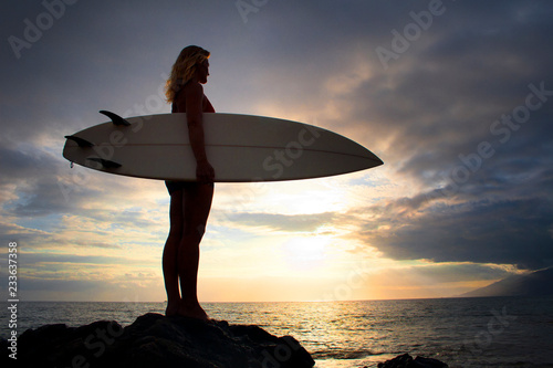 Woman with surfboard standing on rock by sea against cloudy sky during sunset