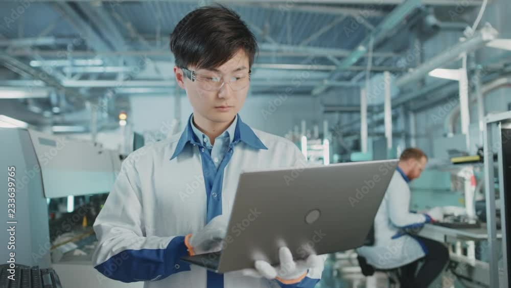 On High-Tech Factory Asian Engineer Uses Laptop Computer for Programming Pick and Place Electronic Machinery for Printed Circuit Board Surface Mount Assembly Line. Production of PCB with SMT Machinery On High-Tech Factory Asian Engineer Uses Laptop Computer for Programming Pick and Place Electronic Machinery for Printed Circuit Board Surface Mount Assembly Line. Production of PCB with SMT Machinery