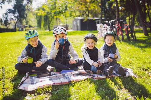 Theme active family holidays nature. group people small little children three brothers and sister sit onblanket near bicycles in park green grass lawn rest and drink drink from cups and thermos tea