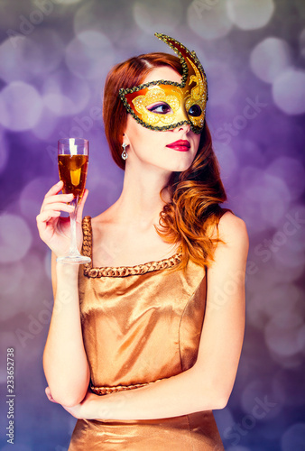 Style redhead girl in golden dress and mask keep glass of drink on gray background with bokeh