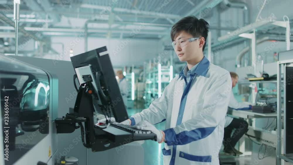 On High Tech Factory Asian Engineer Uses Computer for Programming Pick and Place Surface Mount Electronic Machinery for Printed Circuit Board Assembly Line. On High Tech Factory Asian Engineer Uses Computer for Programming Pick and Place Surface Mount Electronic Machinery for Printed Circuit Board Assembly Line.