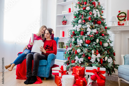 A pretty couple in red sweaters sits on a soft armchair near a tree with gifts in his hands.