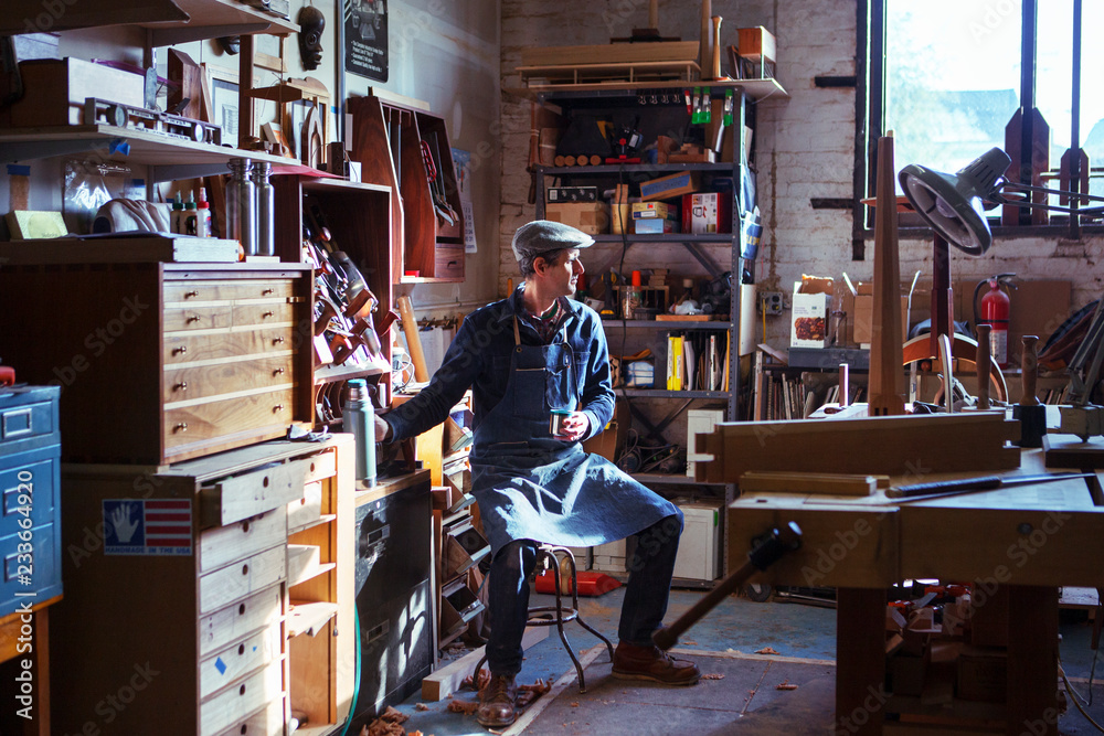 Fototapeta premium Carpenter holding drink looking away while sitting on stool in workshop