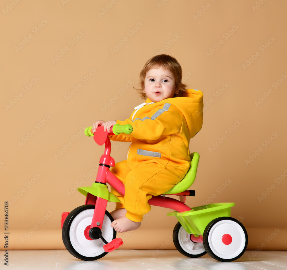 Baby girl kid riding her first bicycle tricycle in warm yellow overalls looking up on light grey Baby girl kid riding her first bicycle tricycle in warm yellow overalls looking up on light grey
