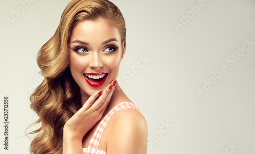 Woman with red lips and nails surprise holds cheeks by hand .Beautiful girl with curly hair surprised and shocked looking on you . Presenting your product. Expressive facial expressions 