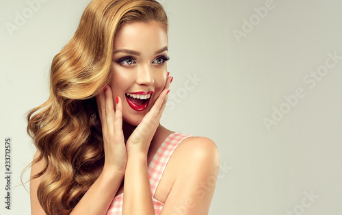 Woman with red lips and nails surprise holds cheeks by hand .Beautiful girl with curly hair surprised and shocked looking on you . Presenting your product. Expressive facial expressions 