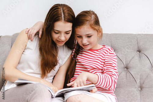 Young woman and her little girl daughter sitting reading books on sofa at home
