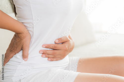 Young woman having painful stomachache and back pain sitting on sofa at home. Chronic gastritis. Abdomen bloating concept.