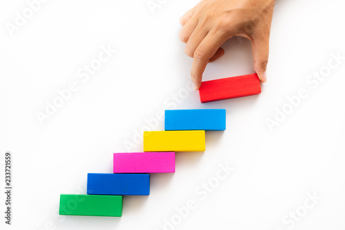 Concept of building success foundation. Women hand put red wooden block on colorful wooden blocks in the shape of a staircase.