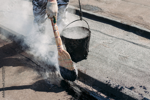 Worker repairs the roof with molten tar from a bucket with a broom.