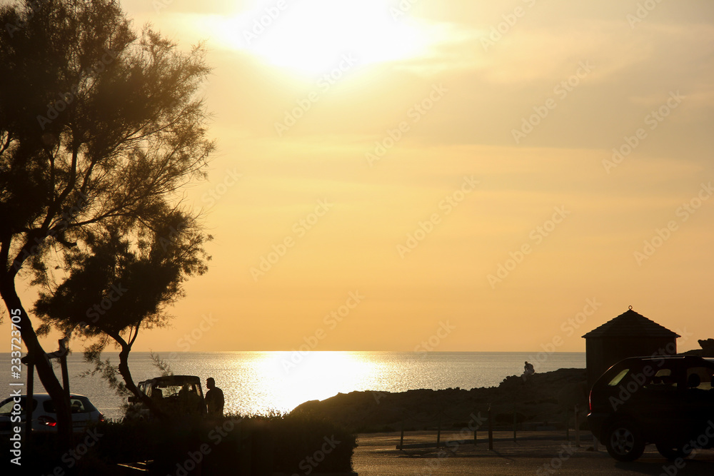 Golden sunset at beautiful seashore. Silhouettes of tree, roof, cars and men on background of sea and sky