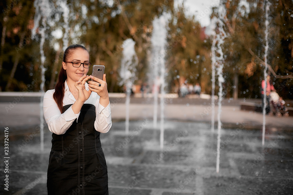 young woman Caucasian appearance in optical glasses in the Park near the fountain with smartphone in hand.