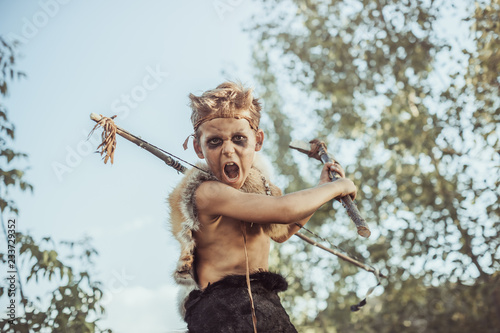 Caveman, manly boy with weapon aggressively shouting. Dramatic action photo of young primitive boy outdoors . Evolution survival concept. Calm boy outside standing in attack pose. Prehistoric tribal