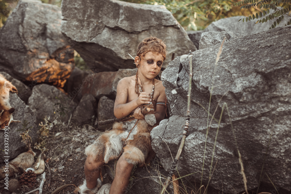 Caveman, manly boy making primitive stone weapon. Funny young primitive boy outdoors near bonfire. Evolution survival concept. Calm boy outside sitting at his rocky settlement. Prehistoric tribal man