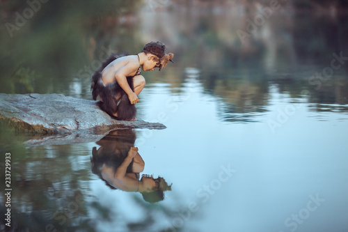 Caveman boy sitting on the rock and looking at him self in the water reflection in lake. Evolution survival concept. Creative art fantasy photo