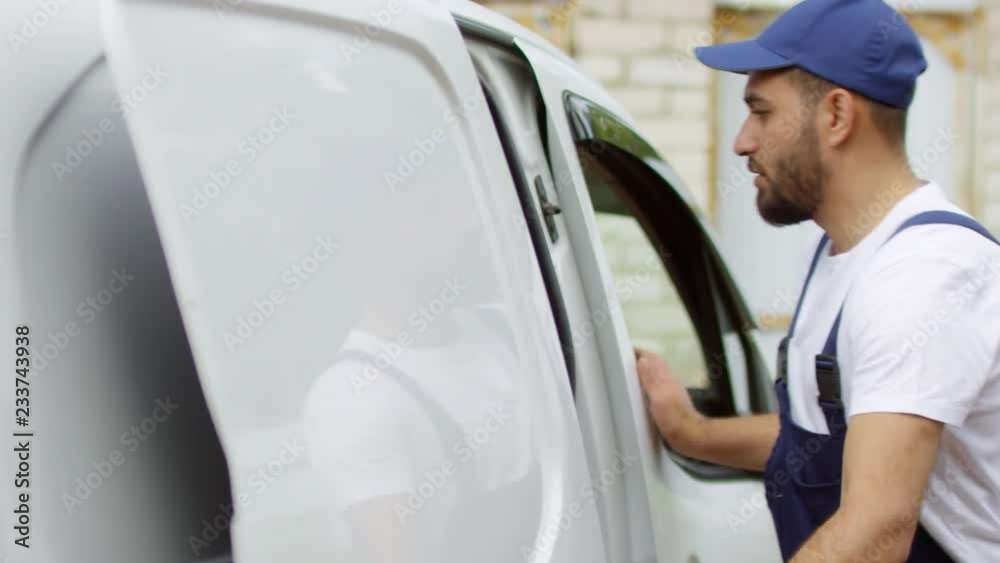 Medium shot of male courier in uniform opening sliding door of white van and taking out package for delivery Medium shot of male courier in uniform opening sliding door of white van and taking out package for delivery