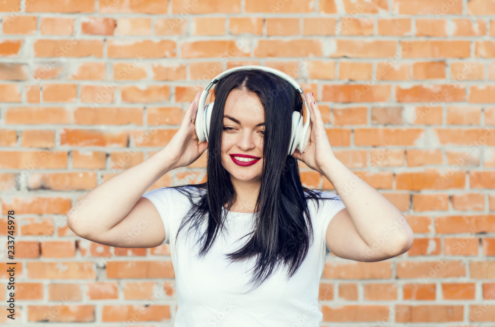 Portrait of a smiling brunette girl on the brick wall background. Listening to music in white headphones. Youth culture, album cover. Portrait of a smiling brunette girl on the brick wall background. Listening to music in white headphones. Youth culture, album cover.
