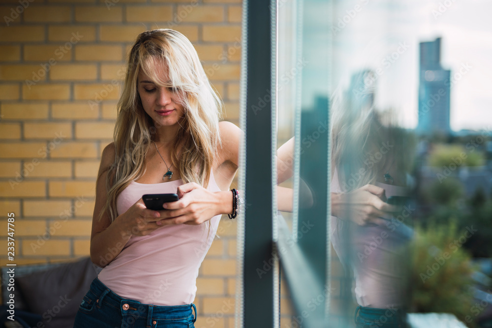 Blond young woman checking cell phone at the window Blond young woman checking cell phone at the window