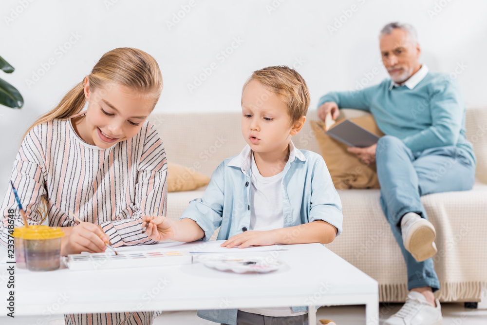 adorable children painting at table while their grandfather reading book behind on sofa at home adorable children painting at table while their grandfather reading book behind on sofa at home