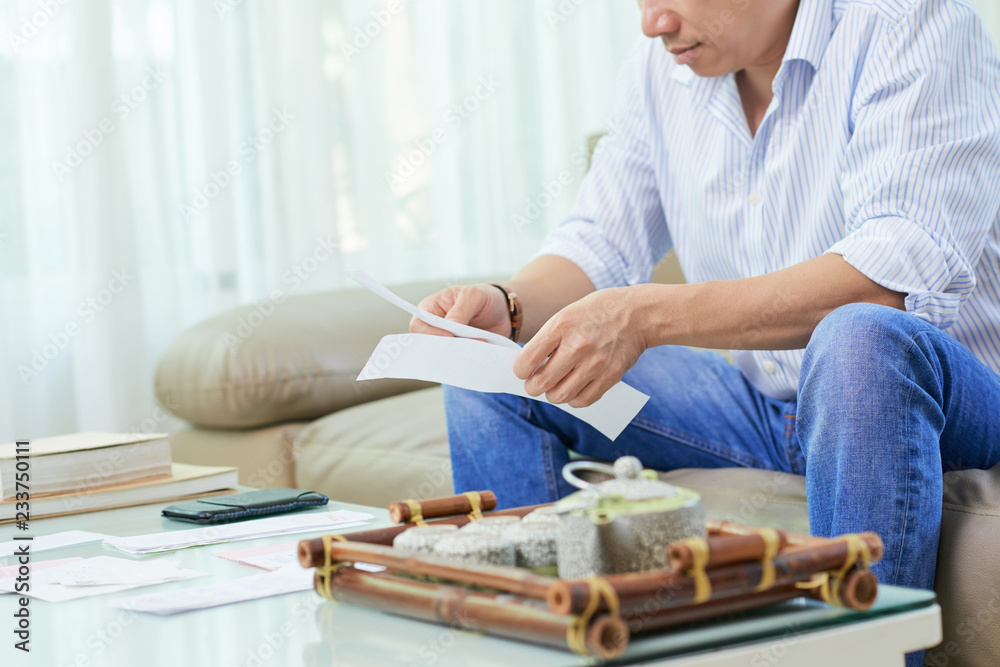 Young man sitting on sofa and checking his unpaid bills or taxes while drinking tea at home Young man sitting on sofa and checking his unpaid bills or taxes while drinking tea at home