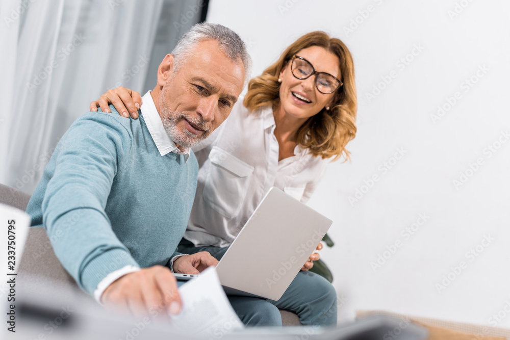 low angle view of man with laptop taking paper from table while his happy wife sitting near at home low angle view of man with laptop taking paper from table while his happy wife sitting near at home