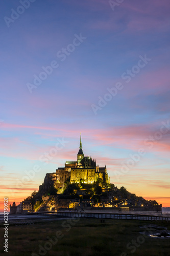 View of the Mont Saint-Michel tidal island, at the limit between Normandy and Brittany in France, illuminated at nightfall with the jetty on stilts and purple clouds in the sky.
