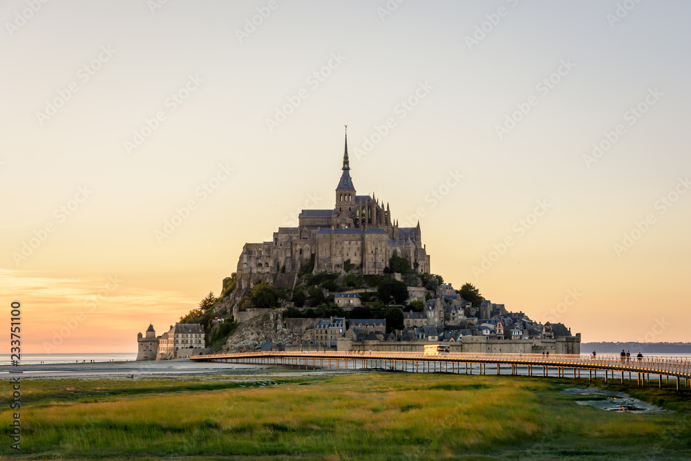 View of the Mont Saint-Michel tidal island at sunset and low tide with a shuttle bus driving on the jetty on stilts, people strolling on the walkway and salt meadows in the foreground.