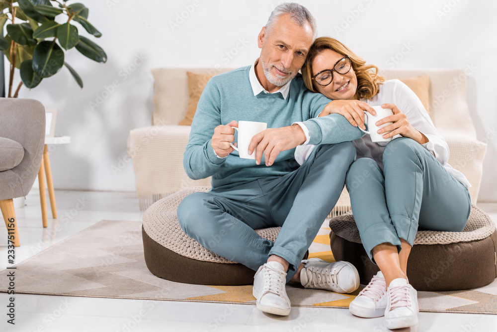 happy mature couple sitting with cups of coffee in living room at home happy mature couple sitting with cups of coffee in living room at home