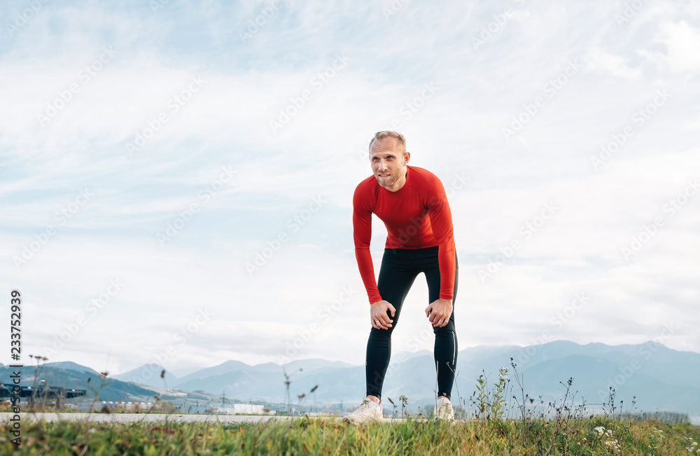 The wide angle shoot of a Man dressed in red long sleeve shirt very tired after the jog  by the road with mountain background and resting to lean forward.