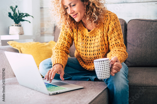 Nice beautiful lady with blonde curly hair work at the notebook sit down on the sofa at home - check on oline shops for cyber monday sales - technology woman concept for alternative office freelance