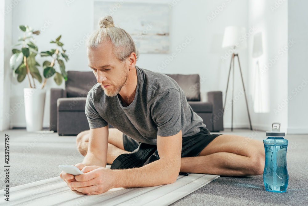sporty adult man sitting on yoga mat and using smartphone at home