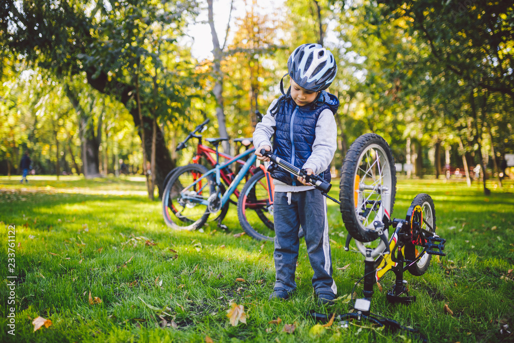 Fototapeta premium Subject child and profession. A little blond Caucasian boy uses a bicycle pump, pumps air into a tire wheel bicycle, a child repairs a bicycle in a meadow in a park in autumn