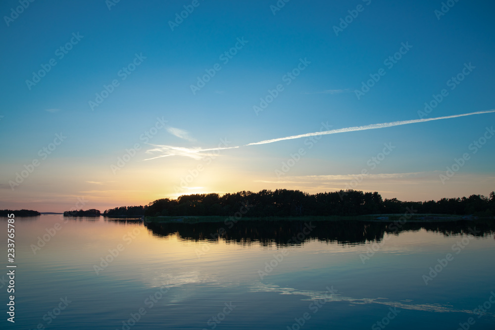 Obraz premium A beautiful and calming sunset accented by a long narrow cloud, a few days after midsummer, reflected on the water of a cove on the Island of Nicklösa in the Åland Islands, Finland.