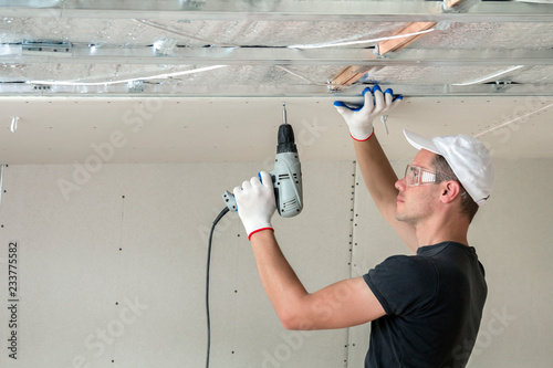 Young man in goggles fixing drywall suspended ceiling to metal frame using electrical screwdriver on ceiling insulated with shiny aluminum foil. Renovation, construction, do it yourself concept. photo