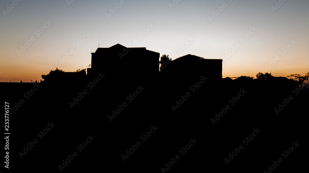 Silhouette of two houses isolated on top of a mountain, while behind them the sun is shining, coloring the sky with red and orange.