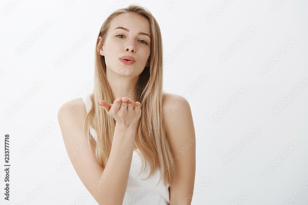 Girl gives encouraging kiss blowing mwah at camera with folded lips and raised hand standing tender and sensual against gray background in casual white tank-top awarding boyfriend for bringing sweets
