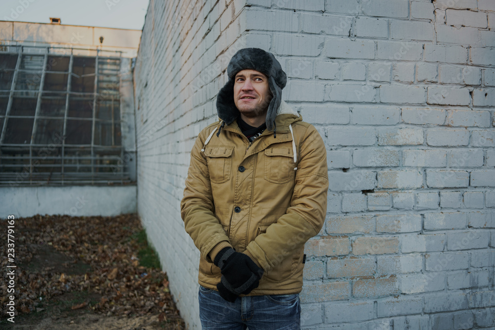 Happy handsome young man in cap with earflaps. The young man in the fur hat. a young guy standing on the street on a cloudy day. emotional portrait of a student. street style